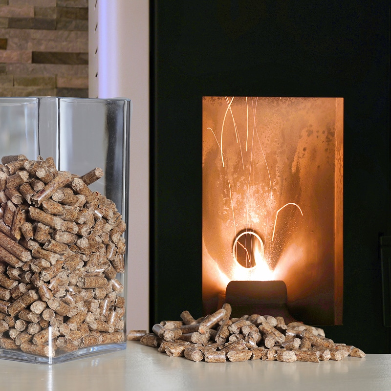 Pellets de bois dans un récipient et éparpillés, à côté d'un poêle à granulés en pleine combustion avec flamme et étincelles.