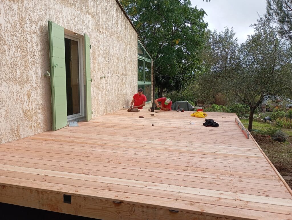Installation terrasse bois : chantier extérieur maison Deux ouvriers installent une nouvelle terrasse en bois clair devant une maison à façade texturée beige et volet vert.