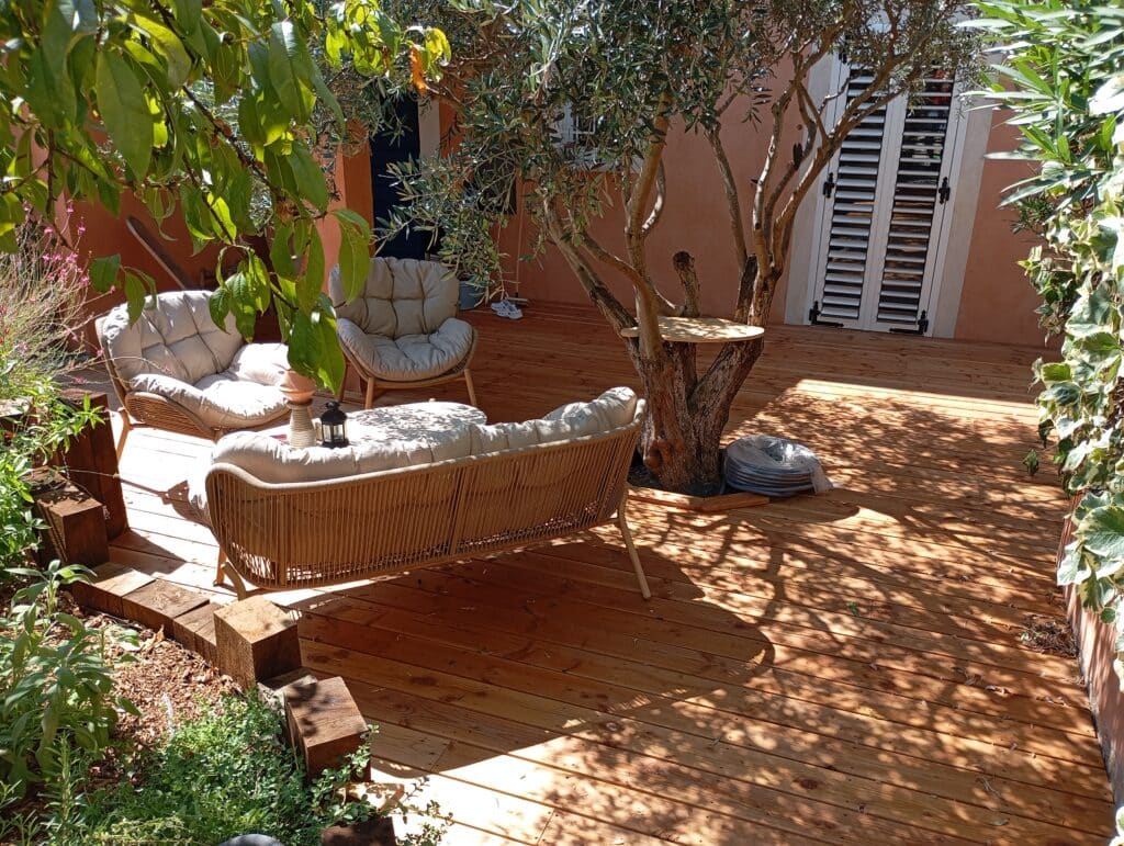 Salon d'extérieur en bois : détente sous l'olivier Terrasse en bois ensoleillée avec mobilier en rotin, sous un olivier et un autre arbre, près d'un mur ocre et volets blancs.
